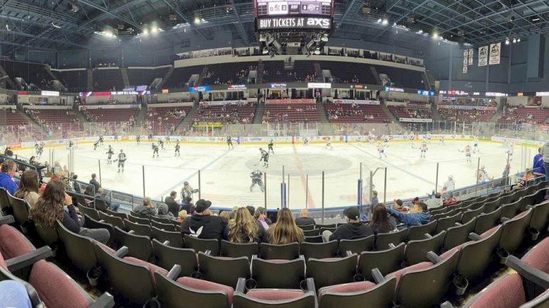 People are watching a hockey game in an indoor arena with players on the ice and a scoreboard above.