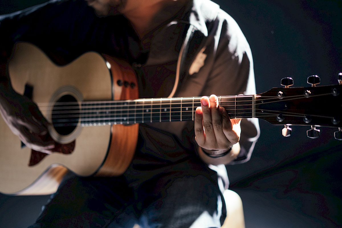 A person is playing an acoustic guitar, with a focus on their hands and the instrument. The background is dimly lit, enhancing the guitar's details.