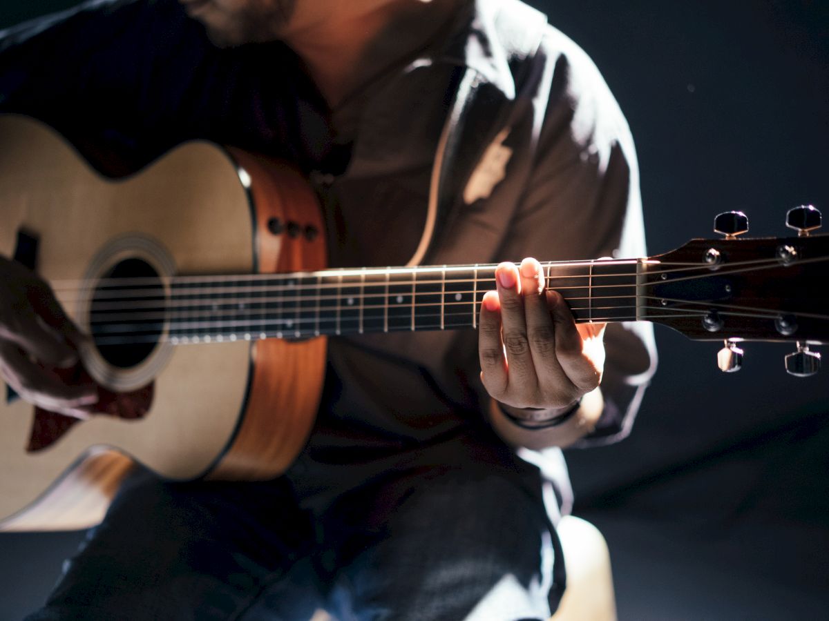 A person is playing an acoustic guitar, with a focus on their hands and the instrument. The background is dimly lit, enhancing the guitar's details.