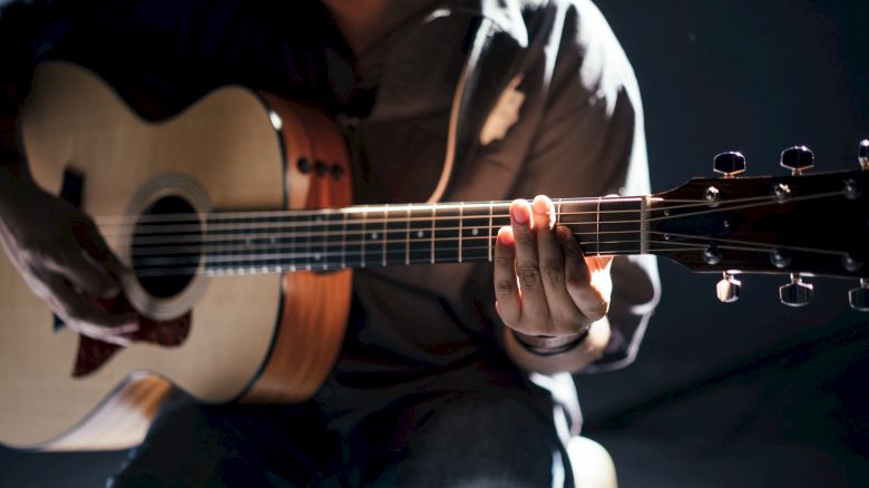 A person is playing an acoustic guitar, with a focus on their hands and the instrument. The background is dimly lit, enhancing the guitar's details.