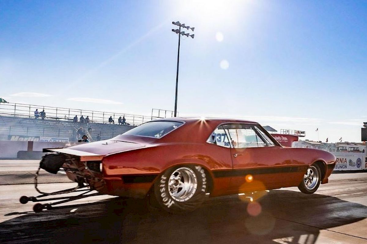 A red muscle car with large rear tires accelerates on a drag strip under bright sunlight, with empty stands and a clear blue sky in the background.