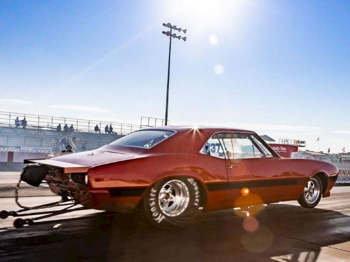 A red muscle car with large rear tires accelerates on a drag strip under bright sunlight, with empty stands and a clear blue sky in the background.