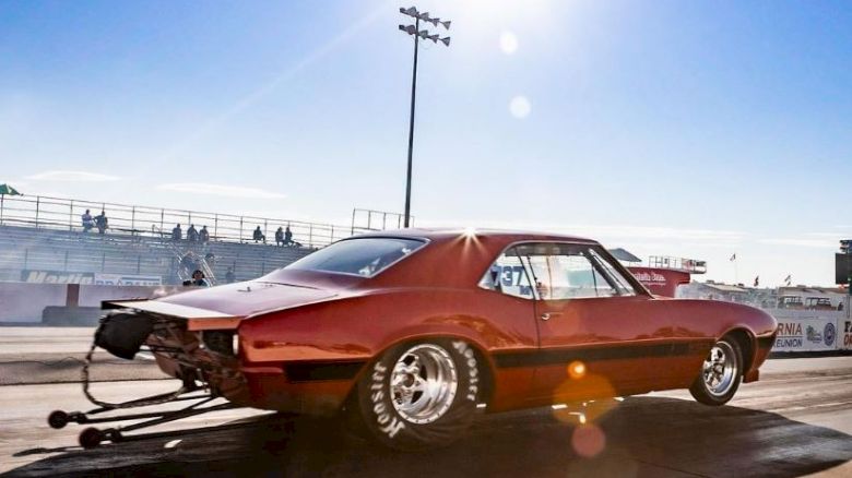 A red muscle car with large rear tires accelerates on a drag strip under bright sunlight, with empty stands and a clear blue sky in the background.