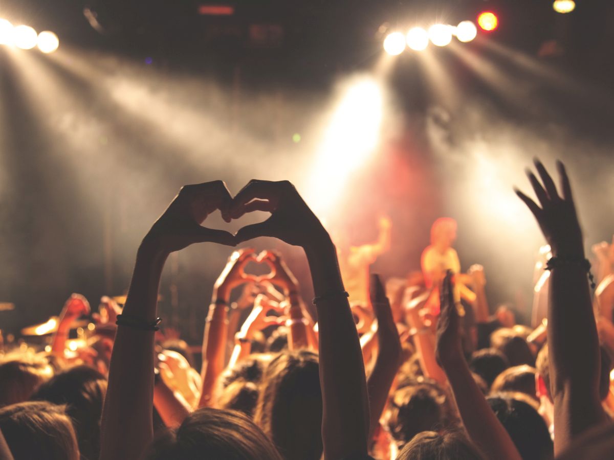 People at a concert are raising their hands, with some forming heart shapes with their fingers, under bright stage lights, creating a lively atmosphere.