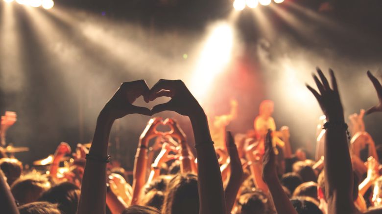 People at a concert are raising their hands, with some forming heart shapes with their fingers, under bright stage lights, creating a lively atmosphere.