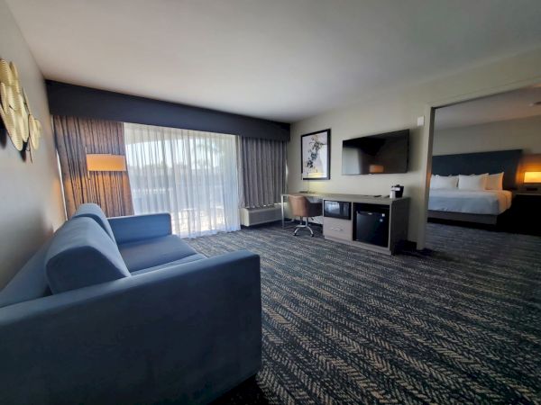 A hotel suite featuring a living area with a sofa, desk, TV, mini-fridge, and a bedroom with a bed, bedside table, lamp, and large window.