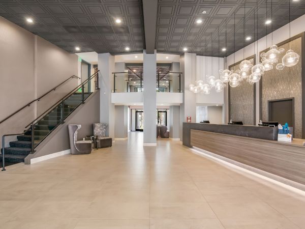 A modern hotel lobby features a reception desk, a staircase, seating areas, elegant lighting, and neutral tones.