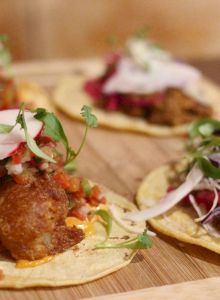 The image shows a wooden board with four tacos, each topped with various ingredients like fish, pico de gallo, radishes, and fresh greens.