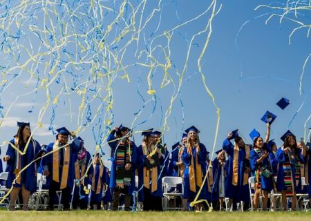 A group of graduates in caps and gowns celebrate outdoors with blue and yellow streamers flying above them under a clear sky, marking graduation day.