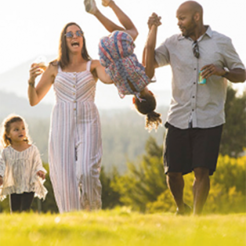 A family of four enjoys an outdoor day, with the parents swinging their child in the air while the other child walks beside them, all smiling.