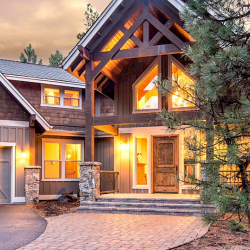 This image features a large, well-lit house with a two-car garage, a wooden facade, and surrounded by tall trees and a stone-paved driveway.