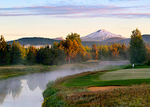 A serene landscape featuring a golf course with a putting green by a reflective river, trees, and a snow-capped mountain in the background.