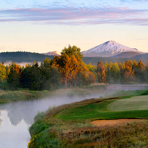 A serene landscape featuring a golf course with a putting green by a reflective river, trees, and a snow-capped mountain in the background.