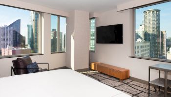 A modern hotel room with large windows showing a cityscape view, a bed, a chair, a wall-mounted TV, and minimalist furniture.