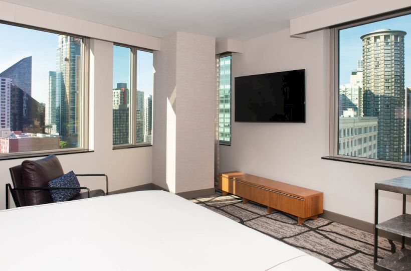 A modern hotel room with large windows showing a cityscape view, a bed, a chair, a wall-mounted TV, and minimalist furniture.