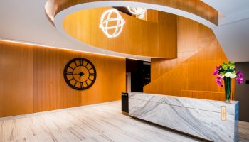 A modern lobby with a curved wooden staircase, marble reception desk, wall clock, and decorative lighting, complemented by a vase of flowers.
