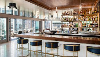 The image shows a modern, well-stocked bar with high stools and glass windows offering a view of city buildings, creating an inviting atmosphere.