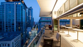 A modern balcony with seating, looking over a cityscape with tall buildings at dusk. The interior shows tables and chairs through large windows.