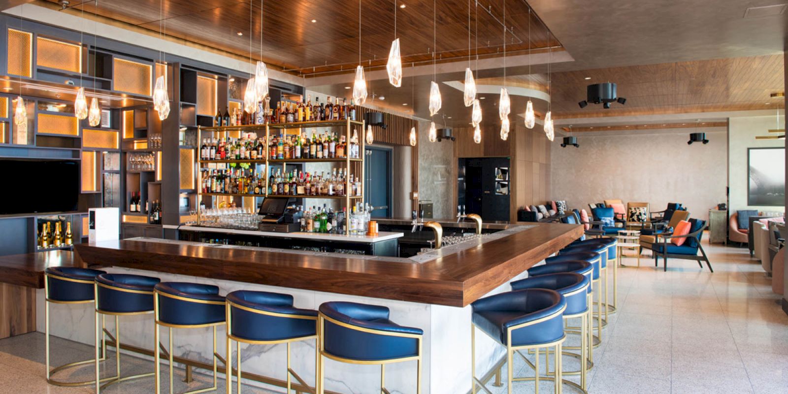 This image shows a modern bar with a stylish counter, blue chairs, and a wide selection of alcohol bottles on shelves. The space is well-lit.