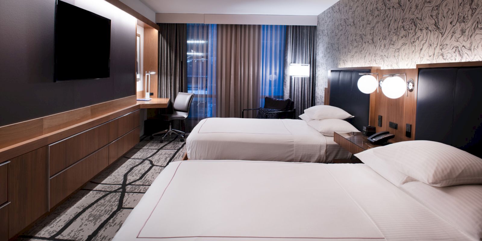 The image shows a modern hotel room with two beds, a desk, a flat-screen TV, and stylish decor, including patterned walls and floor.