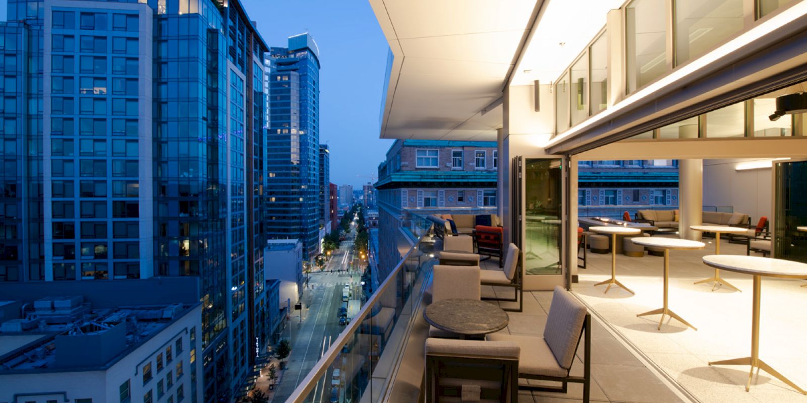 A modern urban balcony at twilight with outdoor seating and tables overlooks city buildings and a street below, with indoor space lit warmly.
