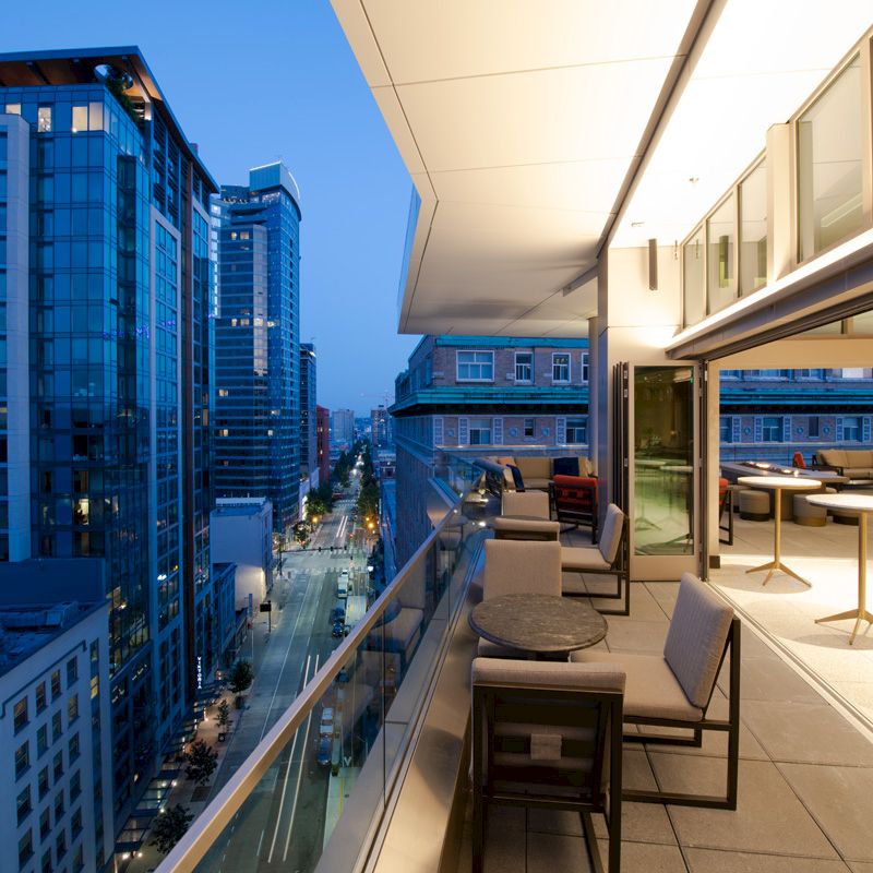 A modern city rooftop terrace at dusk, featuring outdoor seating and tables with a view of illuminated high-rise buildings and a street below.