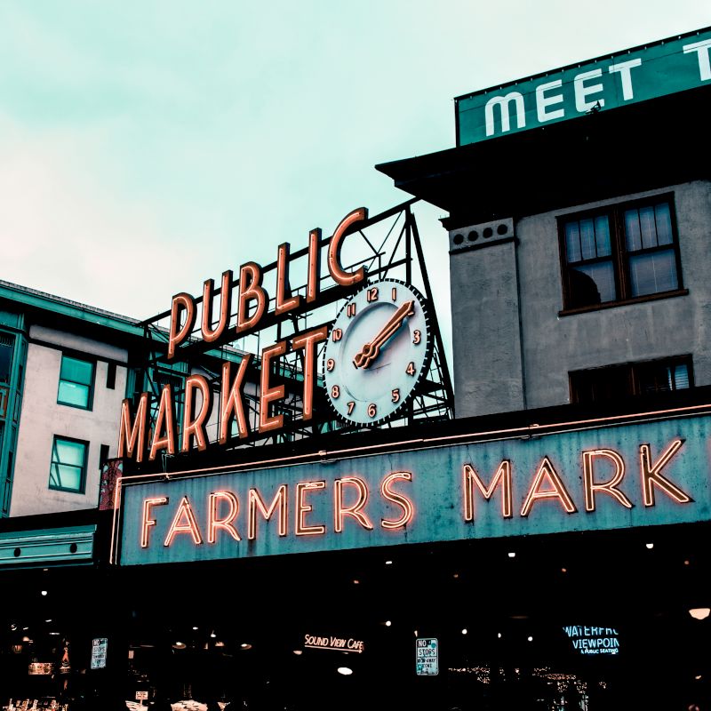 The image shows the iconic signage of a Public Market and Farmers Market with a clock, located among buildings. The sky appears overcast.