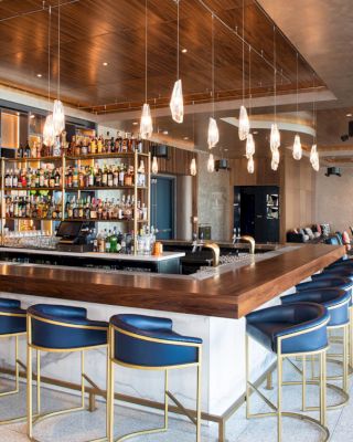 A modern bar with a wooden counter, blue bar stools, and hanging light fixtures. The shelves are stocked with various bottles of alcohol.