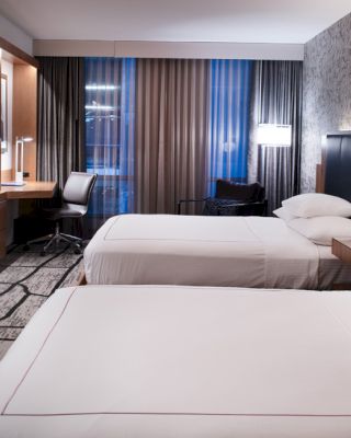 A modern hotel room with two beds, a TV, a desk with a chair, and contemporary decor, including a patterned wall and floor lamp, ending the sentence.