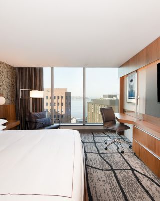 A modern hotel room features a large bed, wall-mounted TV, desk, chair, and a window with a city view. The decor includes wood and artistic wallpaper.