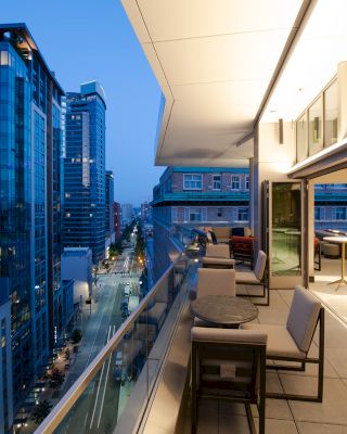 A modern urban balcony at dusk with tables and chairs, overlooking a street lined with tall buildings.