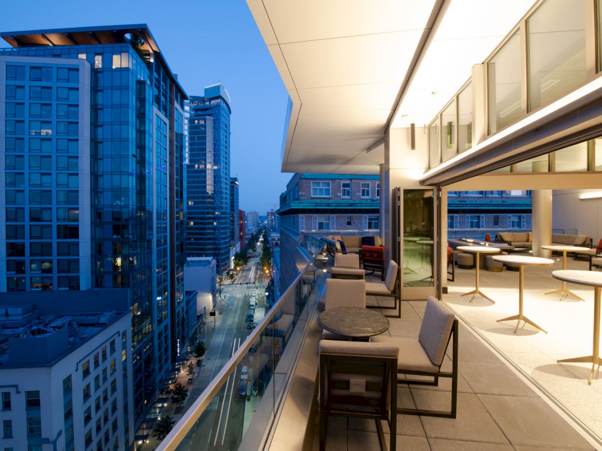 An outdoor balcony of a modern building with seating, tables, and a view of a cityscape with tall buildings at dusk, creating an urban atmosphere.
