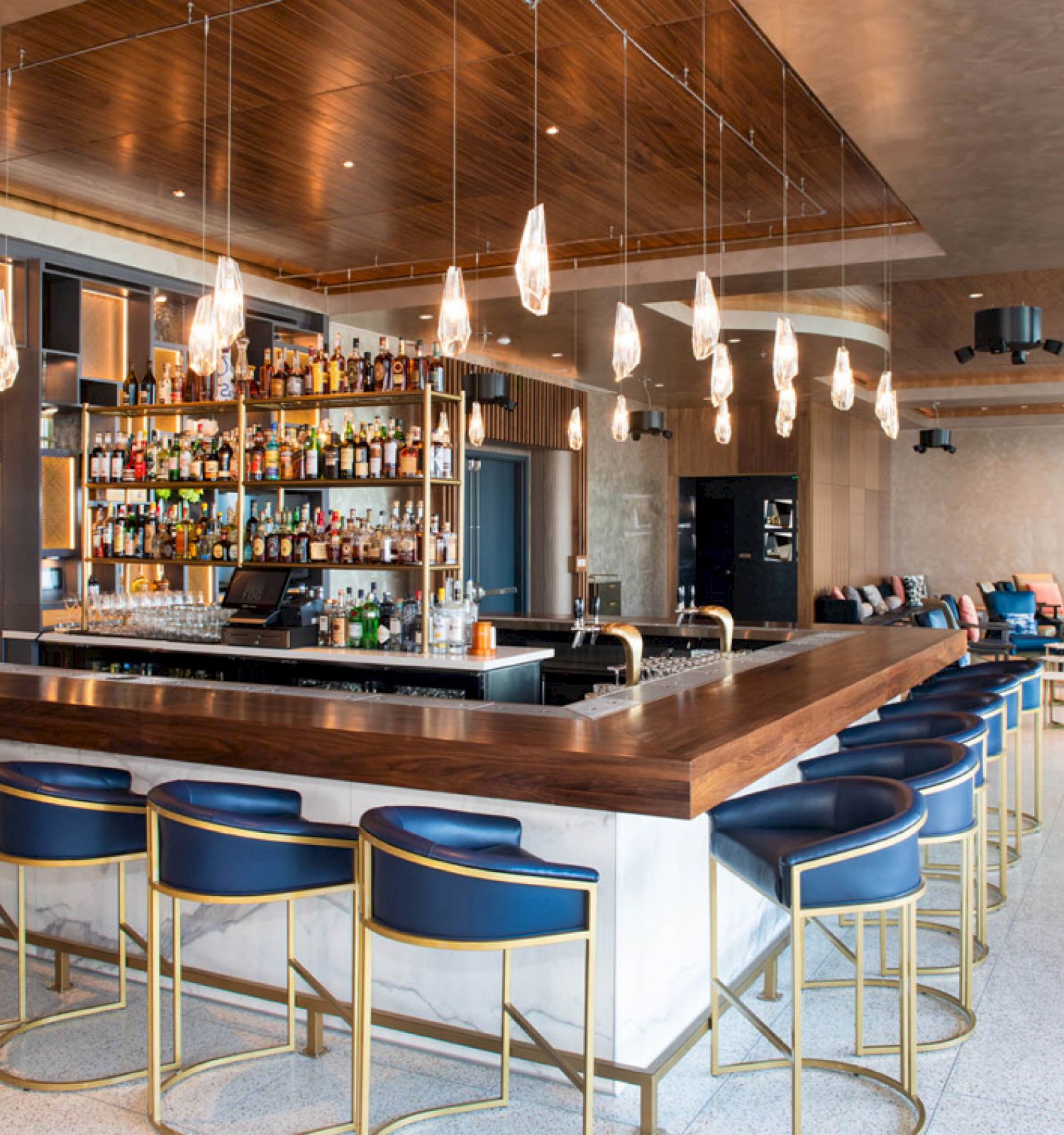 A modern bar with stylish lighting, a well-stocked shelf of bottles, and sleek navy bar stools. There are also several cozy seating areas.