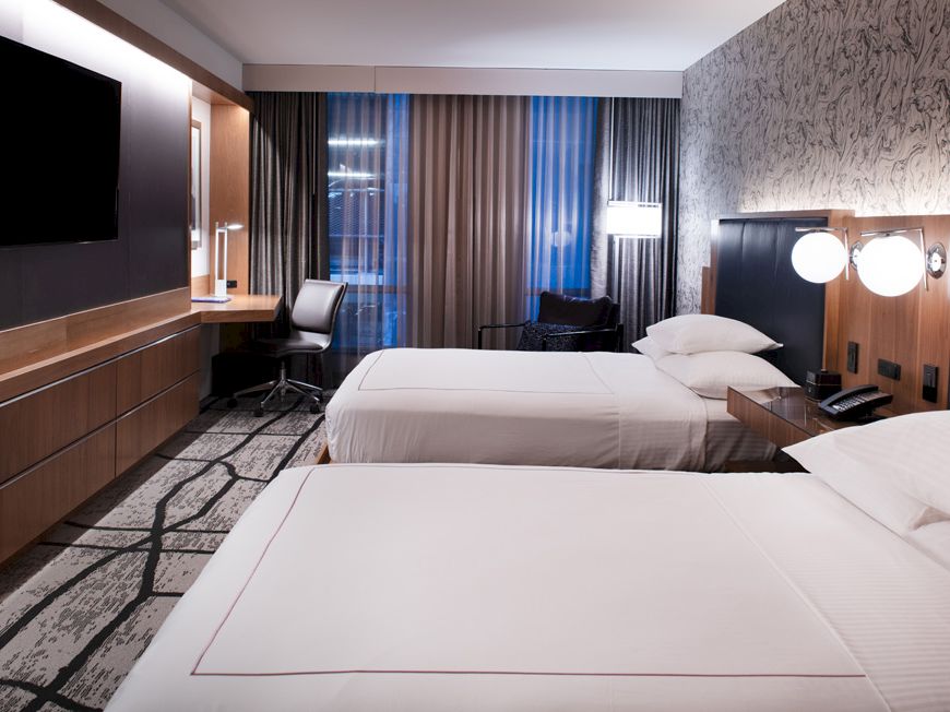 A modern hotel room with two neatly made beds, wall-mounted TV, a desk with a chair by the window, and stylish decor including patterned wallpaper.