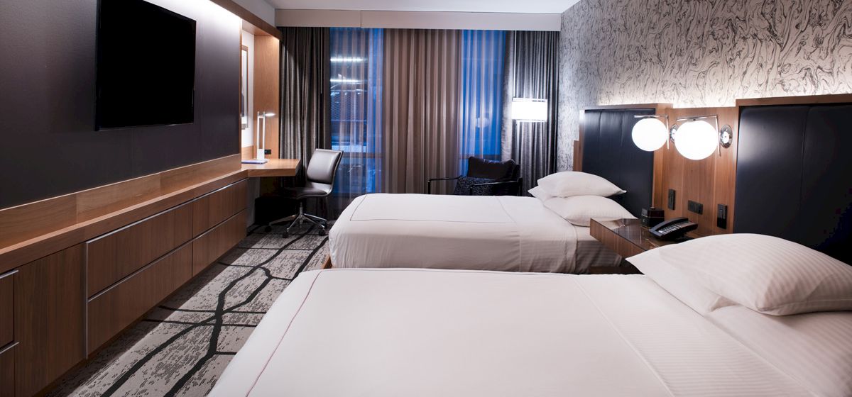 A modern hotel room with two neatly made beds, wall-mounted TV, a desk with a chair by the window, and stylish decor including patterned wallpaper.