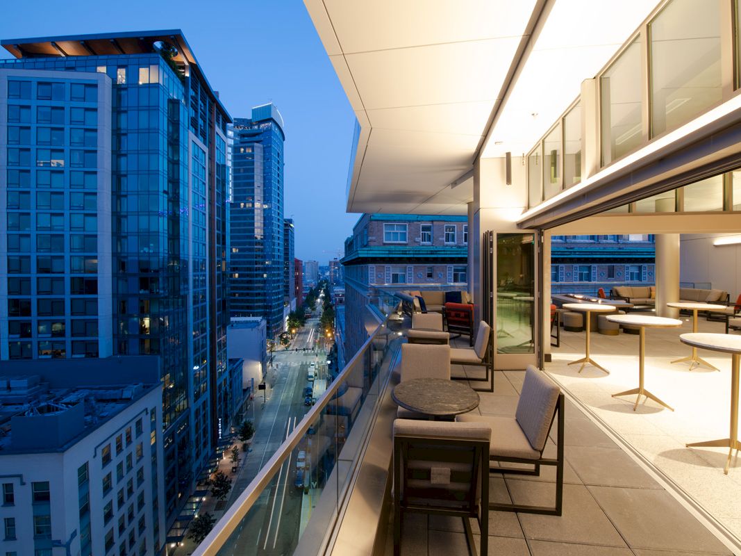 The image shows a modern cityscape at dusk with high-rise buildings, and a stylish balcony featuring seating and tables extending from a commercial space.