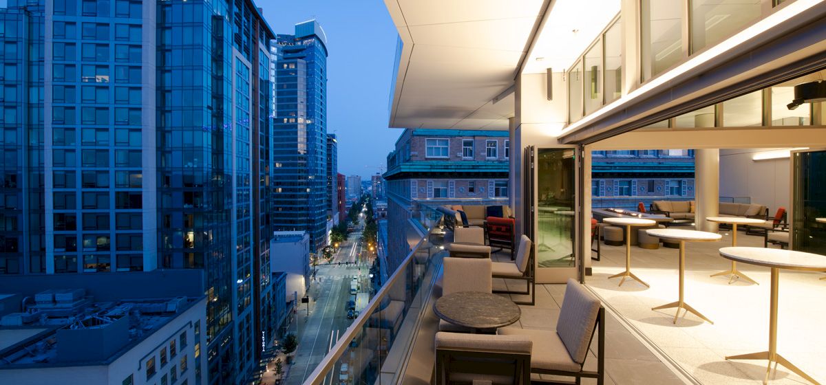 The image shows a modern cityscape at dusk with high-rise buildings, and a stylish balcony featuring seating and tables extending from a commercial space.