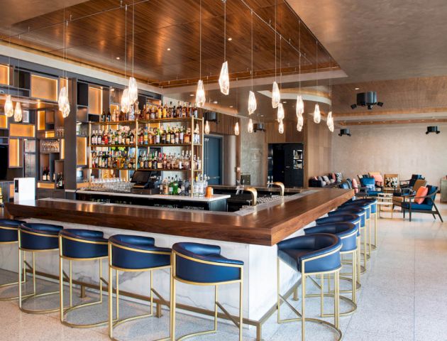 A modern bar with blue barstools, a wooden countertop, hanging lights, and a well-stocked liquor shelf in a spacious, stylishly designed room.