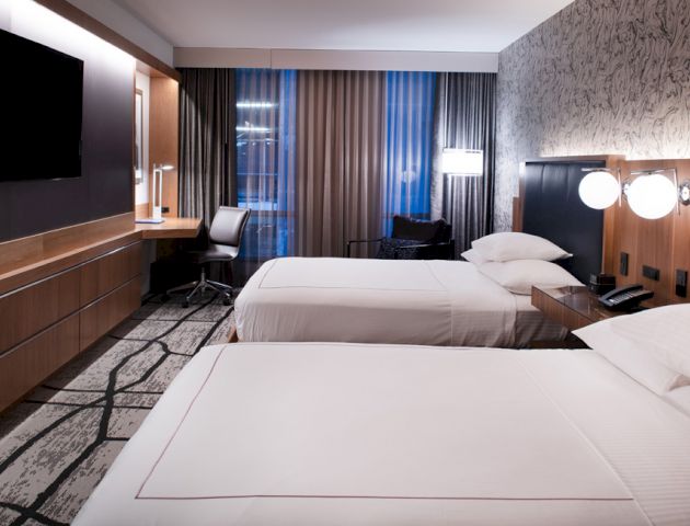 The image shows a modern hotel room with twin beds, a flat-screen TV, a work desk with a chair, and floor-to-ceiling curtains.