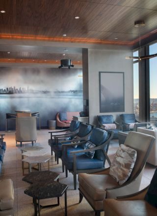 A modern lounge with numerous seats, floor-to-ceiling windows offering a view of the ocean and cityscape, and a mural on the back wall.