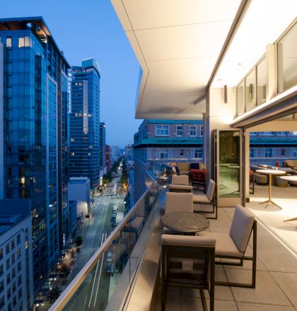 A modern rooftop terrace at dusk with seating and tables, overlooking a city street lined with tall glass buildings.