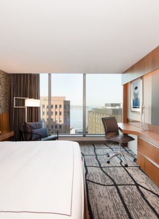 A modern hotel room with a large bed, a desk, a flat-screen TV, and a window offering a city view. The decor is contemporary.