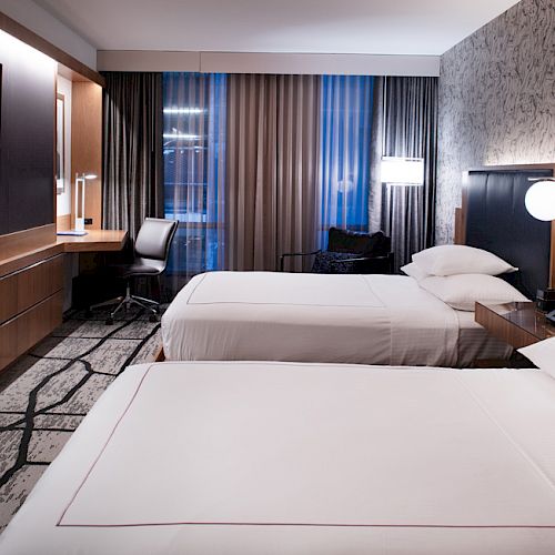 A modern hotel room with two neatly made beds, a desk, chair, TV, and stylish decor, featuring a patterned wall and a large window with drapes.
