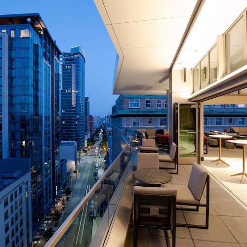 A modern urban balcony at dusk with tables, chairs, and city buildings in the background. The scene captures a blend of indoor and outdoor spaces.