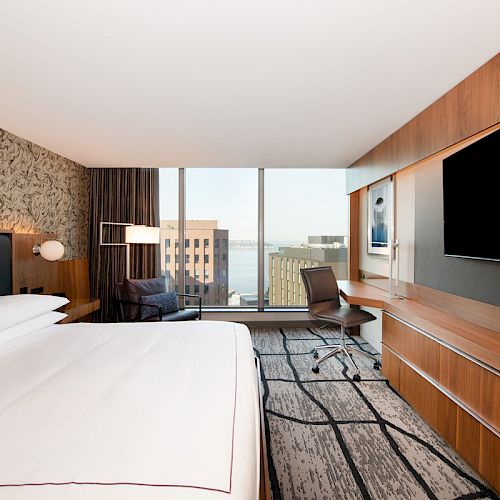 The image shows a modern hotel room with a large bed, a flat-screen TV, a desk and chair, and a window with a city view.