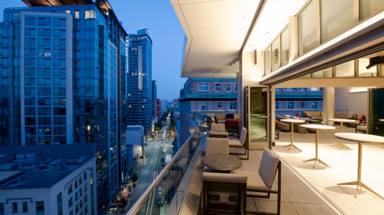 A modern cityscape at dusk with a view from a building's terrace, featuring outdoor seating, high-rise buildings, and a brightly lit street.