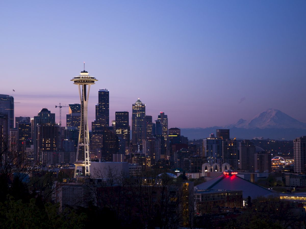 A skyline featuring a prominent needle-like structure, surrounded by illuminated tall buildings, with a mountain in the background at dusk.