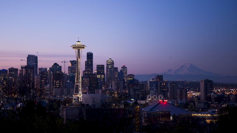 A skyline featuring a prominent needle-like structure, surrounded by illuminated tall buildings, with a mountain in the background at dusk.