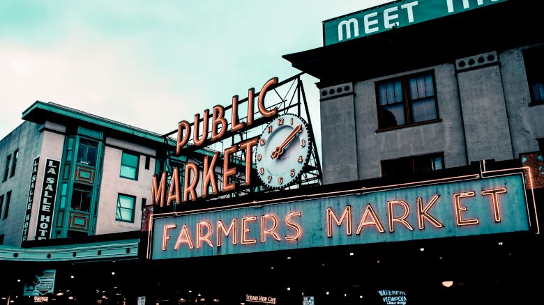 The image shows a neon-lit sign for a "Public Market" and "Farmers Market," with buildings in the background and a visible clock.