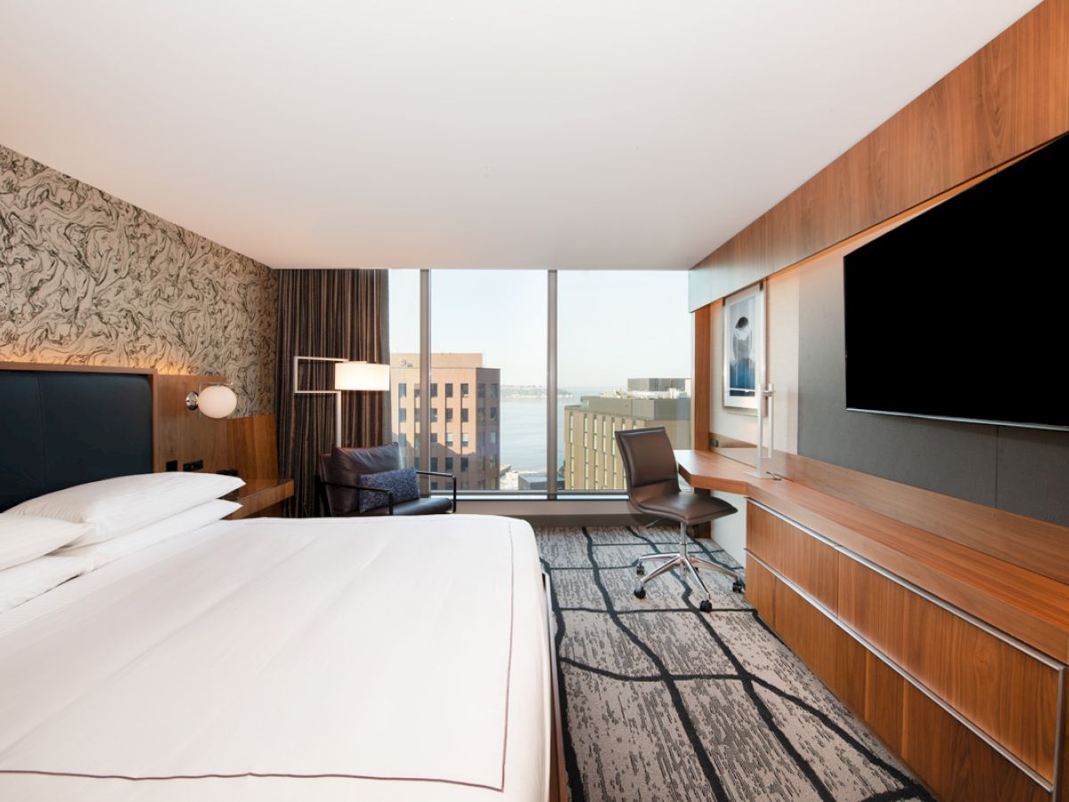 A modern hotel room with a large bed, wall-mounted TV, desk, and floor-to-ceiling windows overlooking buildings and water in the distance.